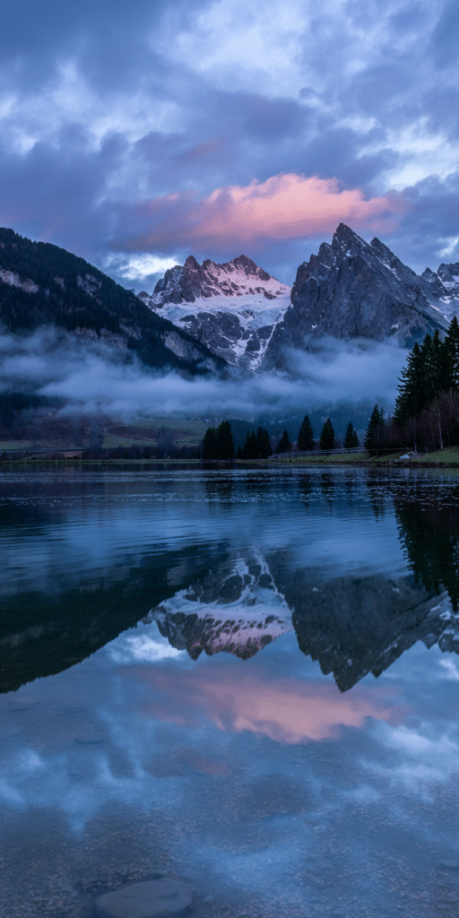 Snow-covered mountains rise above a tranquil lake, where clouds and mist roll across the peaks and reflect in still blue water. The soft pink sunset light contrasts beautifully with the cool tones, making it an ideal wallpaper for those drawn to Beautiful Nature Pictures and Nature Photography. This scenic moment balances power and peace for your lock screen.
