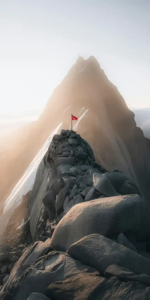 A powerful photo capturing a rocky summit crowned with a red flag, bathed in early morning light. This scene symbolizes ultimate achievement and persistence, making it a standout choice for anyone seeking a Goal Wallpaper Motivation or building a Successful Life Vision Board. The light, texture, and symbolism evoke strength, independence, and clarity of purpose—ideal for iPhones as a daily boost.
